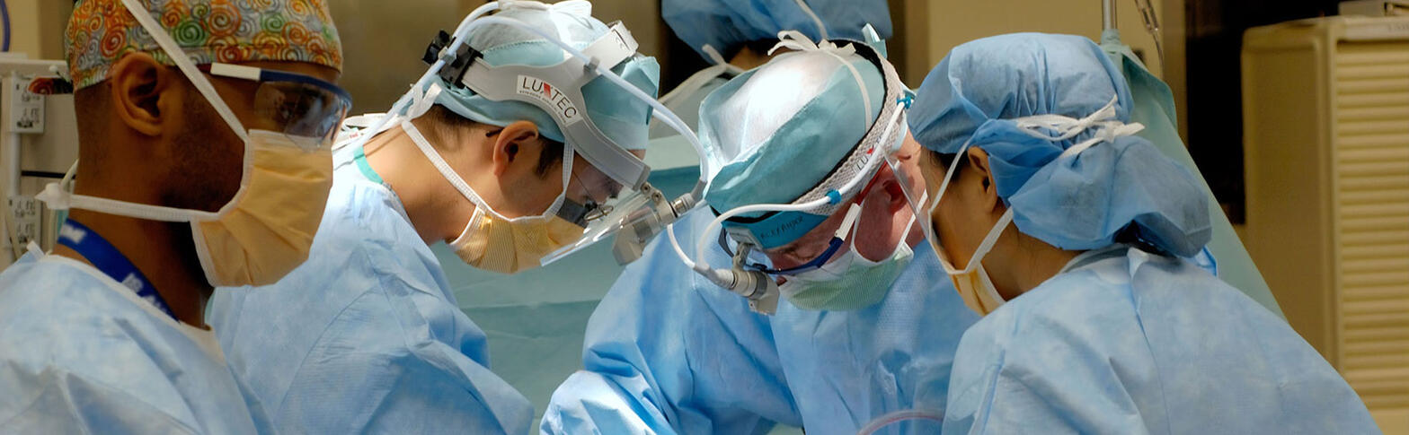 Surgeons dressed in medical gowns, masks, caps, and glasses lean over an operating table. 