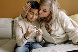 Adult wearing beige sweater hugging child wearing beige sweater, seated together on sofa. Child using vision white board and finger stim device. 