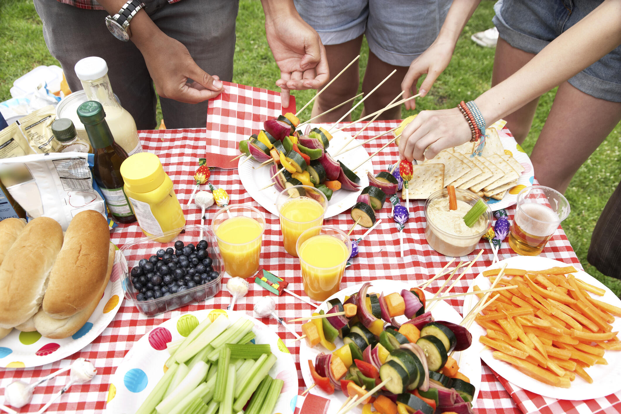 A table full of color foods for a picnic. People's hands reaching for food.