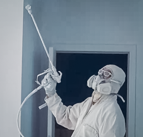 A worker wearing a respirator and goggles sprays paint on an interior wall of a house.
