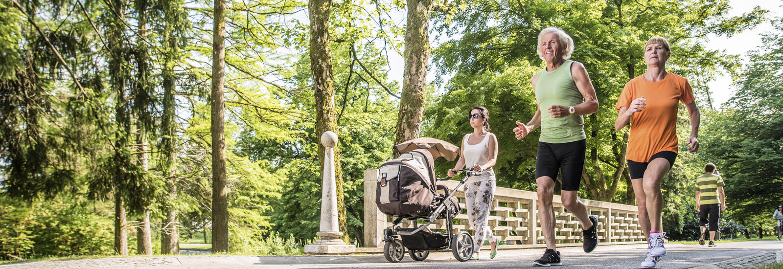 Two people jogging and one person walking with a stroller in tree-lined park.