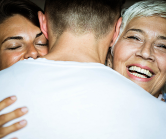 Man in white shirt being hugged by a woman with brown hair and another woman with white hair.